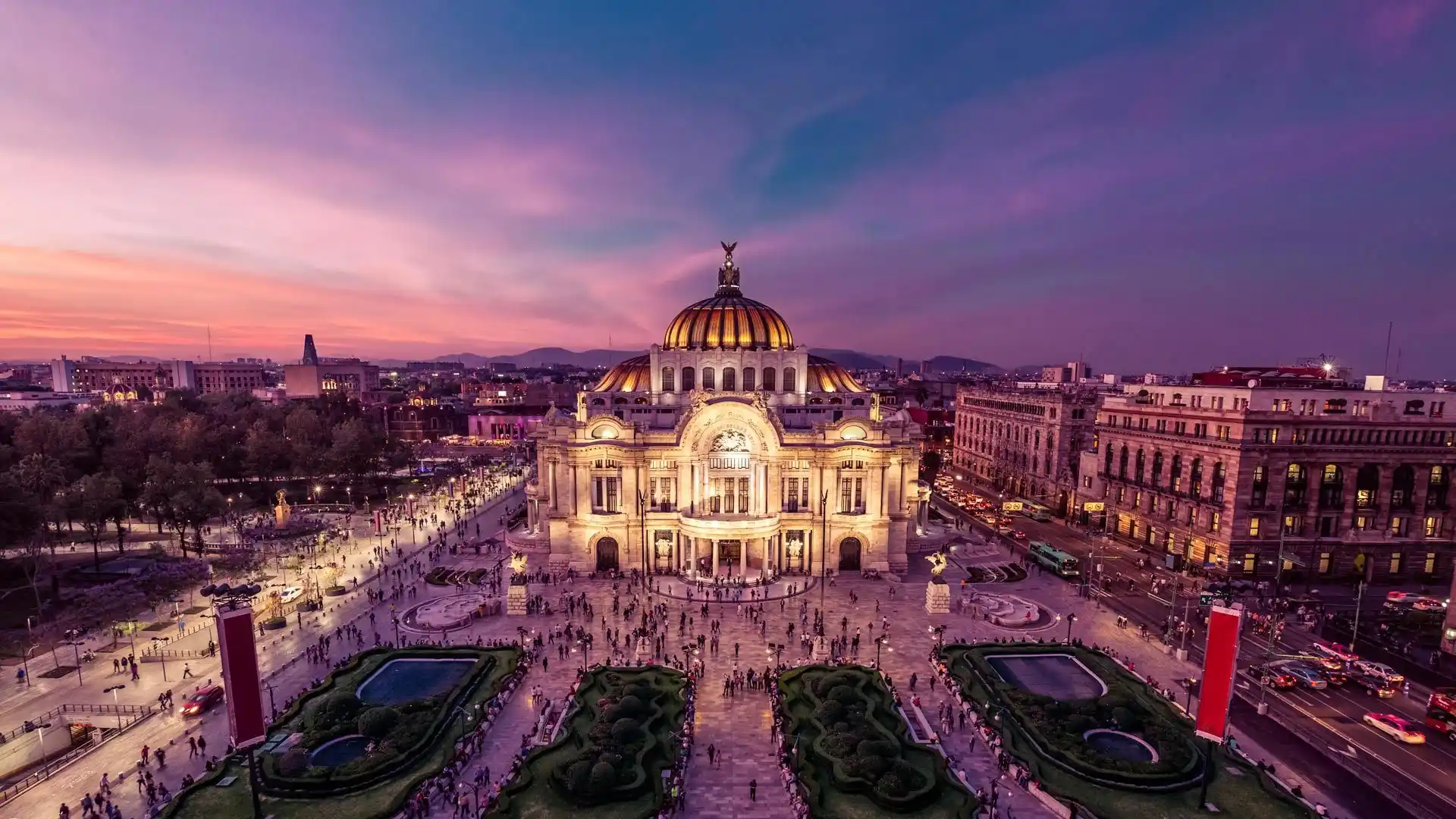 Aerial view of the Palacio de Bellas Artes in Mexico City at dusk. The grand, domed building is illuminated with a warm glow, surrounded by bustling streets and a beautifully landscaped garden. The sky is painted in hues of pink and purple as the sun sets over Mexico City.