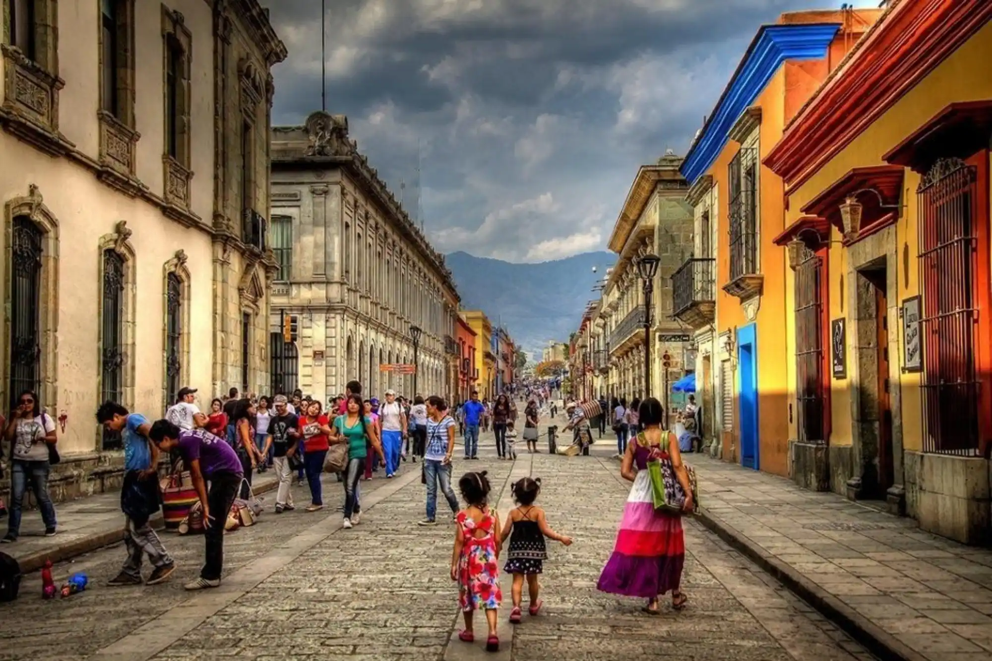 A vibrant street scene in Oaxaca City showcases colorful buildings lining both sides. People are walking, conversing, and engaging in various activities. Two young children in bright dresses hold hands in the foreground. Mountains are visible in the distance under a cloudy sky.