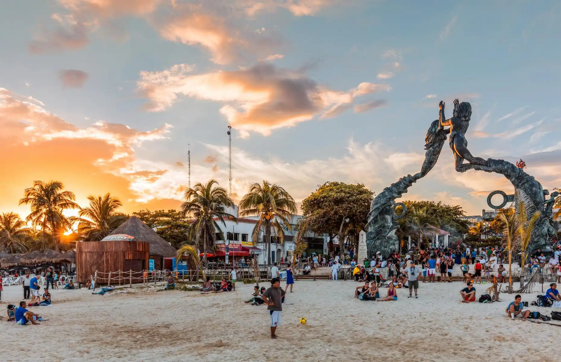 A busy beach scene at sunset with groups of people enjoying the sandy shore in Playa del Carmen. In the background, tall palm trees and thatched huts create a tropical atmosphere. A large, arched sculpture with two human figures reaching toward each other is a focal point.