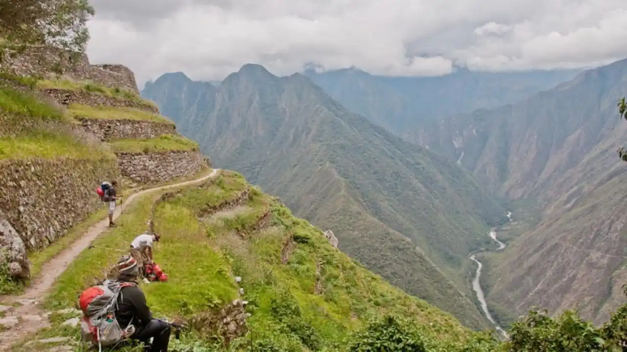 Traveling to Inca Trail Itinerary: How to Get There and What to Pack 2 A scenic view of terraced green hills and a winding path in the foreground, with hikers walking and resting. A river flows through the lush valley below, surrounded by tall, mist-covered mountains under a cloudy sky, reminiscent of trekking the historic Inca Trails.