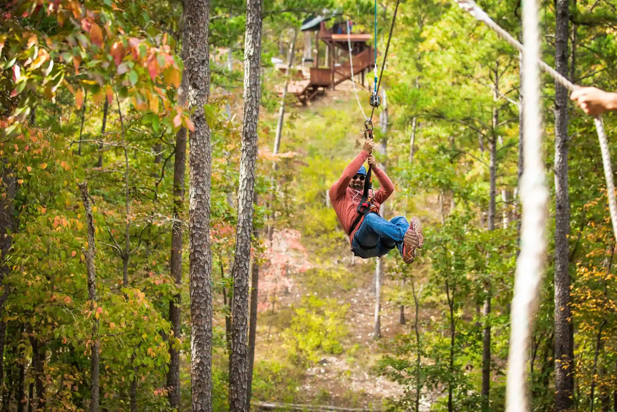 The Ultimate Summer Guide for Solo Travelers 12 A person wearing a red jacket and blue jeans is zip-lining through a lush forest with green and autumn-colored foliage at Rugaru Adventures in Broken Bow, OK. A wooden cabin is visible in the background amid the trees. The person appears to be having an exciting adventure.