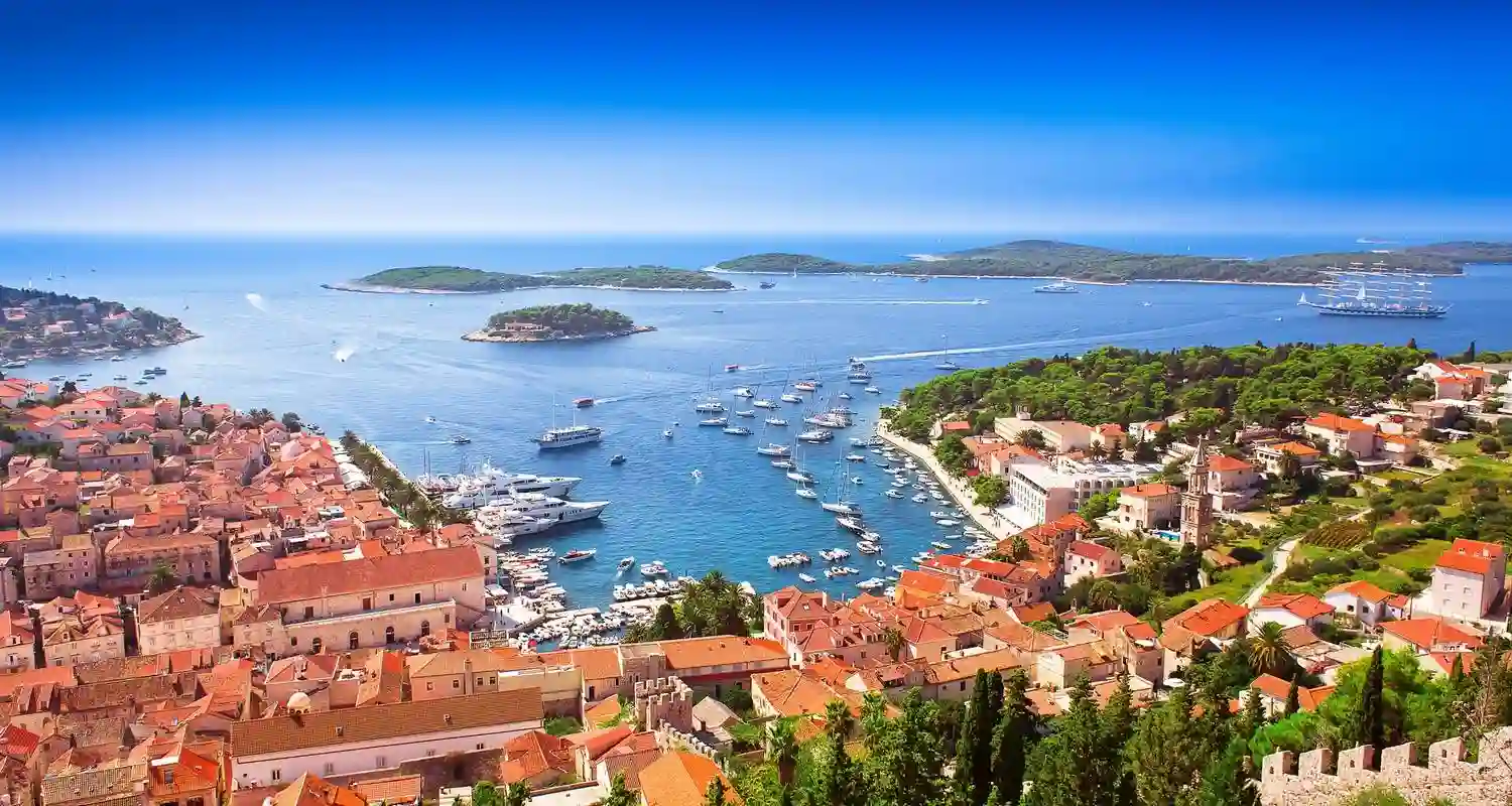 A scenic view of a coastal town with clay-tiled rooftops, a turquoise bay filled with boats, and several green islands in the distance under a clear blue sky.