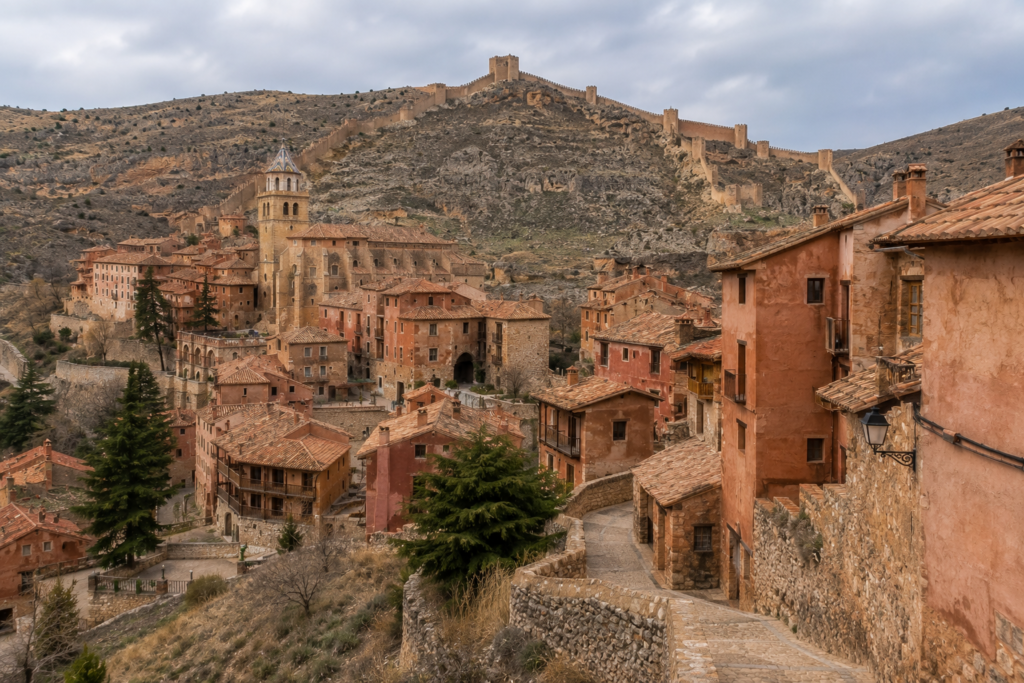 Hidden Gems in Europe - Albarracín, Spain medieval hillside village with terracotta rooftops, ancient stone walls, and rugged mountains under a cloudy sky.