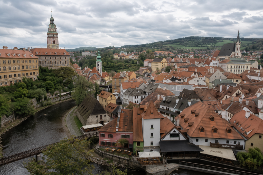 Hidden Gems in Europe - Český Krumlov, Czech Republic panoramic view of the medieval town with red rooftops, winding river, historic castle, and church tower beneath cloudy skies.