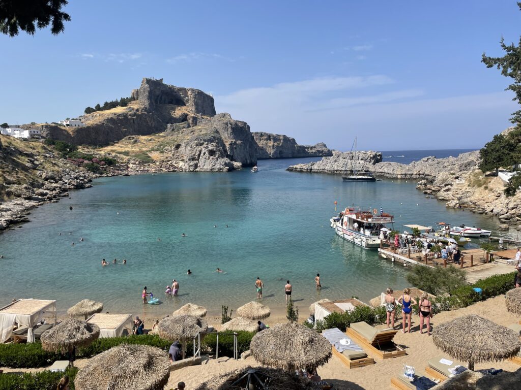 Tourists relaxing and swimming in the crystal-clear waters of St Paul’s Bay below the Acropolis of Lindos in Rhodes, Greece.