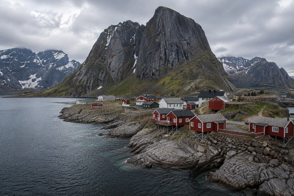 Hidden Gems in Europe - Lofoten Islands, Norway dramatic fjord landscape with red fishing cabins, rugged mountains, rocky shoreline, and calm Arctic waters beneath overcast skies.