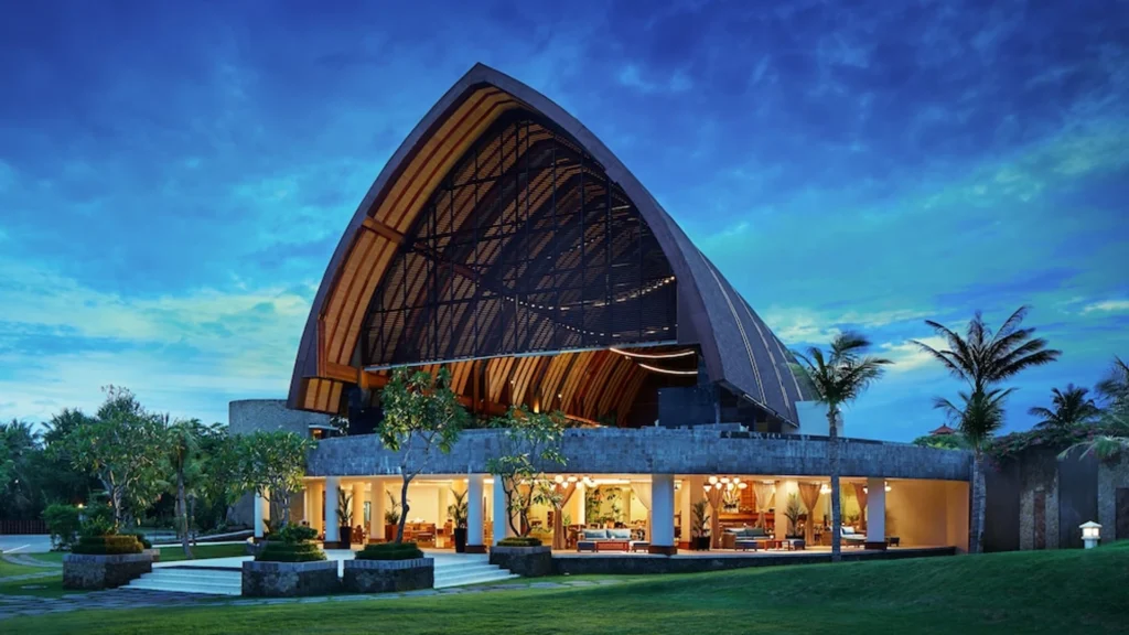 Exterior view of MERUSAKA Nusa Dua resort in Bali featuring dramatic Balinese-inspired architecture illuminated at sunset with tropical gardens and open-air lobby.