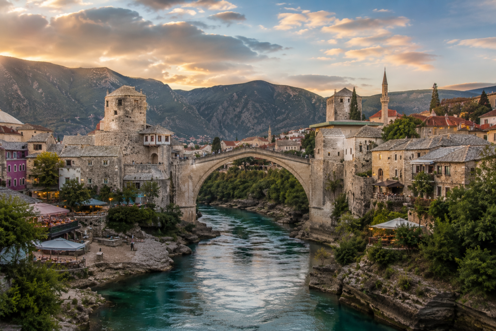Hidden Gems in Europe - Mostar, Bosnia and Herzegovina historic stone bridge arching over a turquoise river with Ottoman-style buildings, rocky cliffs, and mountain scenery in warm evening light.