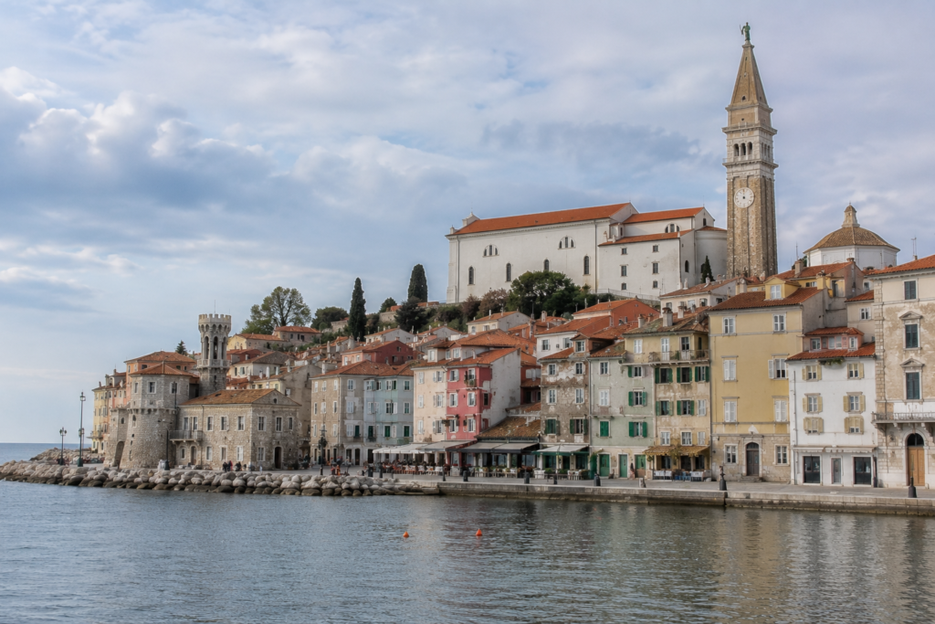 Hidden Gems in Europe - Piran, Slovenia seaside town with Venetian-style buildings, terracotta rooftops, stone bell tower, and Adriatic coastline under soft cloudy skies.