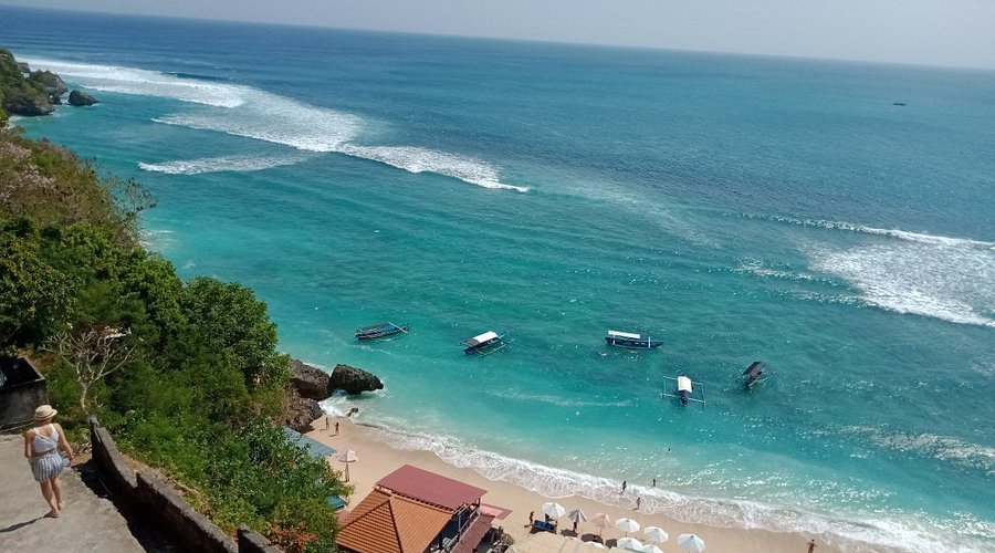 Elevated view of Padang Padang Beach in Bali, with turquoise water, rolling white waves, several small boats near the sandy shore, and lush green vegetation along the cliffside.