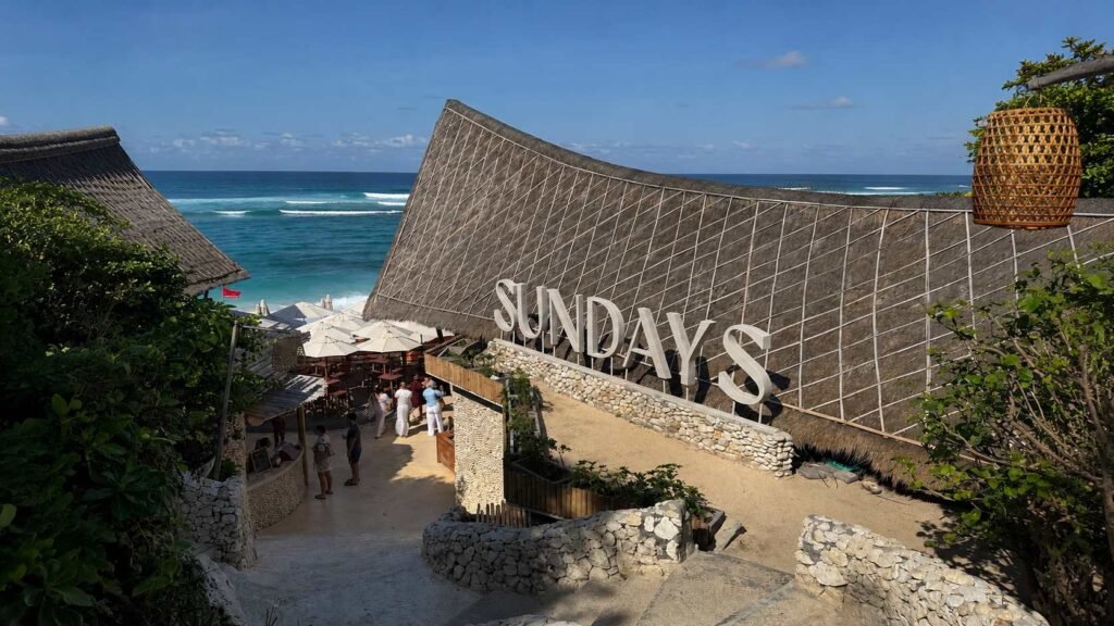 Entrance to Sundays Beach Club in Uluwatu, Bali, with a large thatched-roof building, stone pathways and greenery overlooking the bright blue ocean.